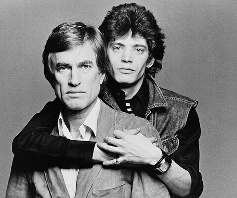 A black-and-white studio portrait photograph of two White men, one standing behind the other with his arms wrapped around the other's shoulders.