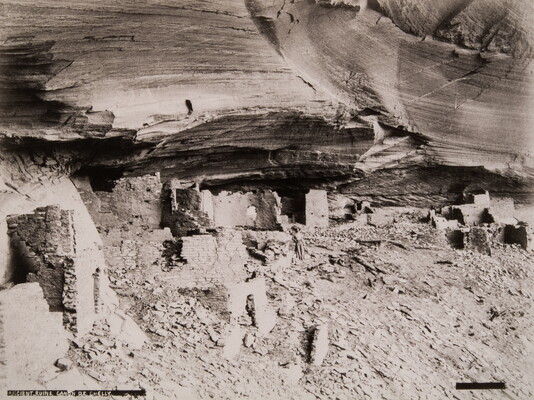 A black-and-white photograph of adobe ruins under an overhanging cliff ledge.
