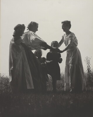 A black-and-white photograph of White women and children wearing long dresses holding hands to form a circle in a grassy field.