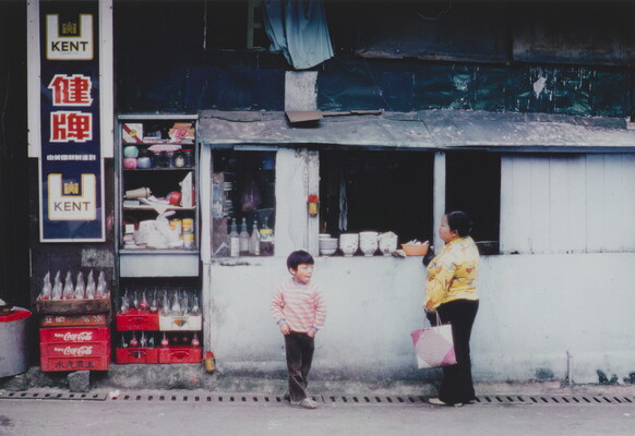A color photograph of an Asian woman and young child standing at the window of a small white building that looks like it serves food.
