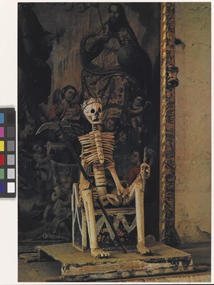 A color photograph of a skeleton seated on a throne, wearing a crown and holding a scythe in one hand, in front of a painting.