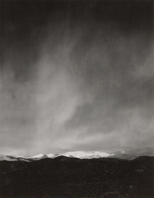 A black-and-white photograph of a snow-covered mountain range with a dark storm forming in the sky.