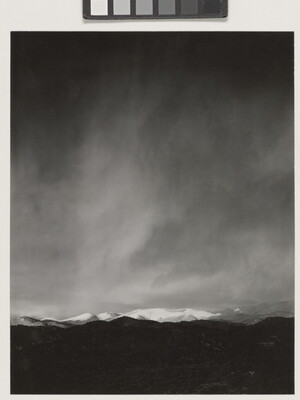 A black-and-white photograph of a snow-covered mountain range with a dark storm forming in the sky.