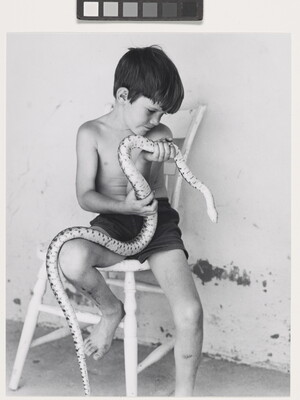 A black-and-white photograph of a shirtless young White boy sitting on a wooden chair holding a large snake.