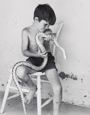 A black-and-white photograph of a shirtless young White boy sitting on a wooden chair holding a large snake.