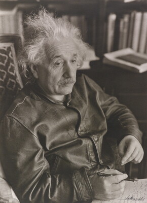 A black-and-white photograph of a seated older White man with shaggy hair and a mustache wearing a leather jacket; notepad on his lap.