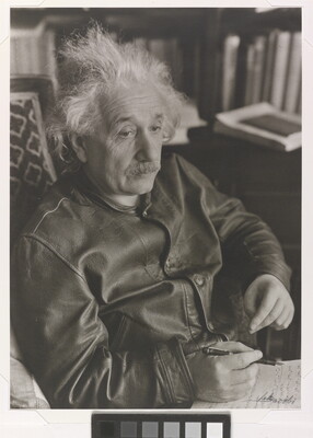 A black-and-white photograph of a seated older White man with shaggy hair and a mustache wearing a leather jacket; notepad on his lap.