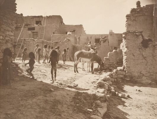 A black-and-white photograph of Indigenous children playing in a courtyard surrounded by a multi-level pueblo as donkeys graze nearby.