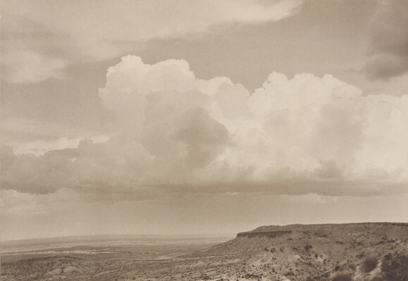 A black-and-white photograph of a mesa with a plain below it beneath a cloudy sky.
