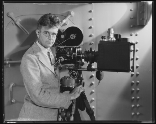 A black-and-white photograph of a White man wearing granny glasses and a light-colored suit standing next to an old movie camera.