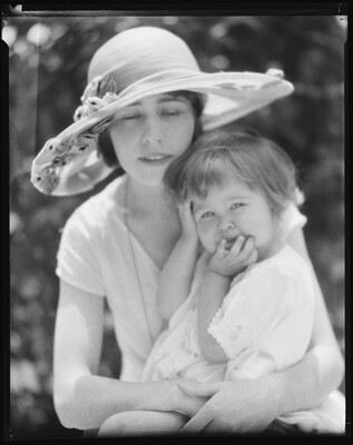 A black-and-white image of a White woman wearing a large sunhat holding a child on her lap, both wearing white.