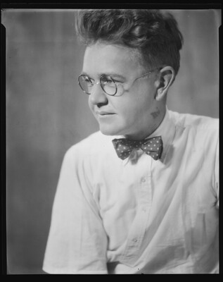 A black-and-white image of a young White man wearing round wire-rimmed glasses, a polka dot bow tie, and a white button-down shirt.