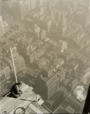 A black-and-white photograph taken from above of a man kneeling precariously on the edge of a construction platform suspended high over a city skyline.