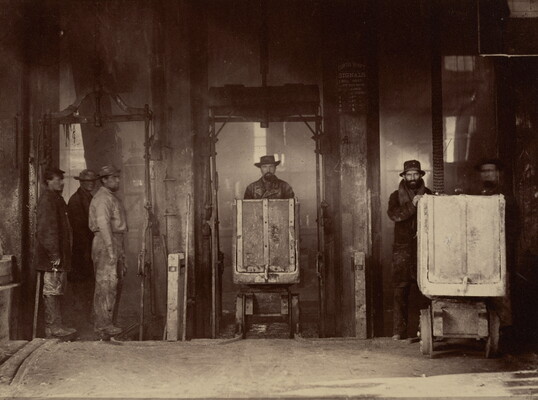 A sepia-toned photograph of men pushing minecarts towards the viewer in an industrial setting with beams of light back-lighting them.