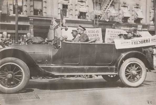 A black-and-white photograph of a convertible vehicle on an urban street with four Black people in the car, a banner draped on the side, and signs displayed in the background.