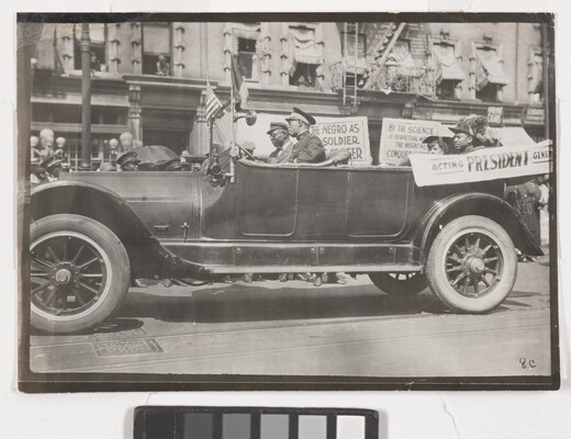 A black-and-white photograph of a convertible vehicle on an urban street with four Black people in the car, a banner draped on the side, and signs displayed in the background.
