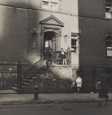 A black-and-white photograph of the entrance to an apartment building with children playing on the outside of the railing on the steps that lead to the door.