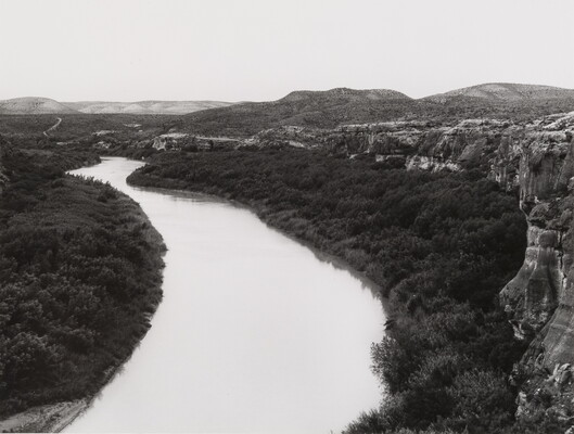 A black-and-white photograph of a river surrounded by many mountains, trees, and rock formations.