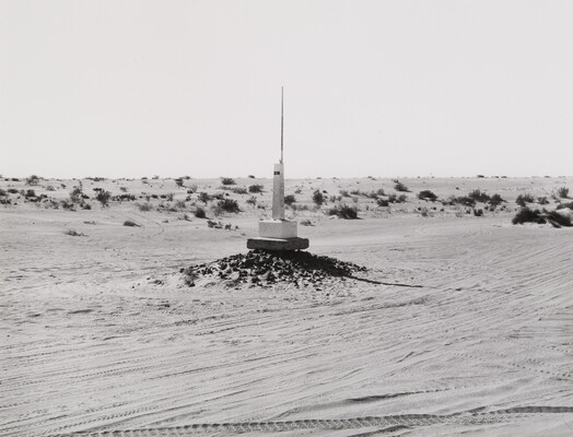 A black-and-white photograph of a white obelisk on a mound of rocks in the center of a flat, sandy landscape with scrub brush in the distance.