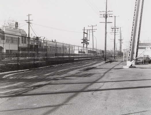 A black-and-white photograph of a tall wire fence with buildings on one side, a flat lot with steel girders on the other.