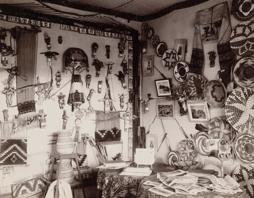 A black-and-white photograph of an interior room, the walls covered with Native American dolls, baskets, blankets, and pottery.