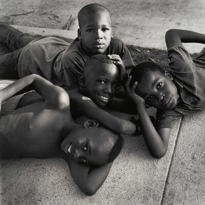 A black-and-white photograph of four young Black boys lying on each other and a sidewalk, smiling up at the viewer.