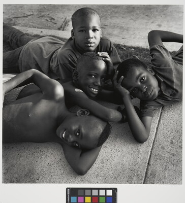 A black-and-white photograph of four young Black boys lying on each other and a sidewalk, smiling up at the viewer.