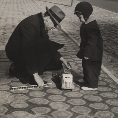 A black-and-white photograph of a small boy bundled in winter clothing watching as a man in a fedora crouches down to reach toward a toy fire truck at the boy's feet.