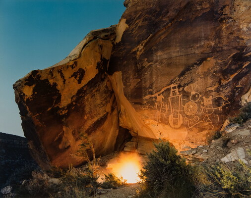 A color photograph of small campfire illuminating a rock wall with several human-like petroglyphs on it.