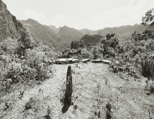 A black-and-white photograph of a single column of stone standing on an overlook lined with stones at the edge; trees and mountains in the distance.