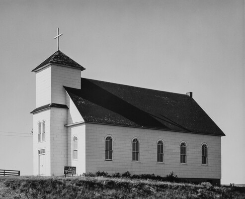 A black-and-white photograph of a small, white clapboard church, viewed from the side, under clear skies.