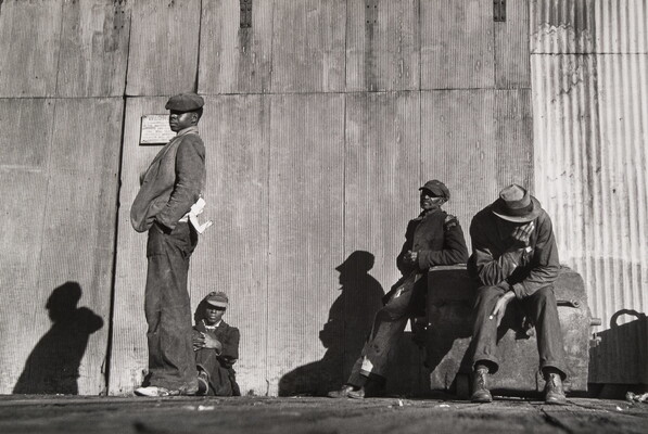 A black-and-white photograph of four Black men, three sitting and one standing, all wearing hats in front of a corrugated metal wall.