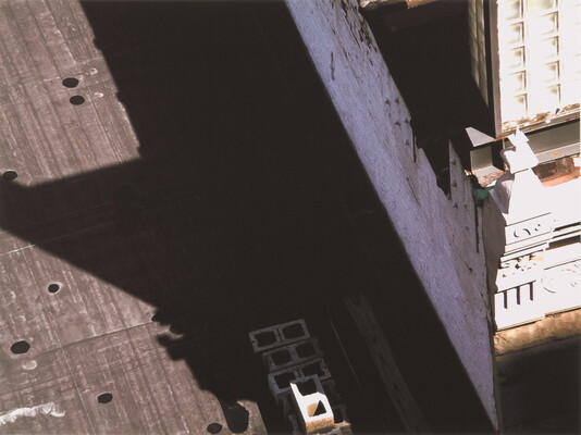 A black-and-white photograph of a building and the shadow it casts across the top of a neighboring building as viewed from the above.