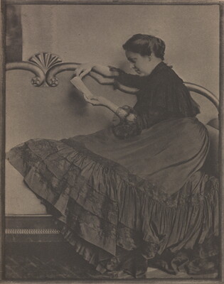 A black-and-white photograph of a seated White woman in profile, hair pulled up and wearing a long ruffled dress, reading on an ornate Victorian settee.