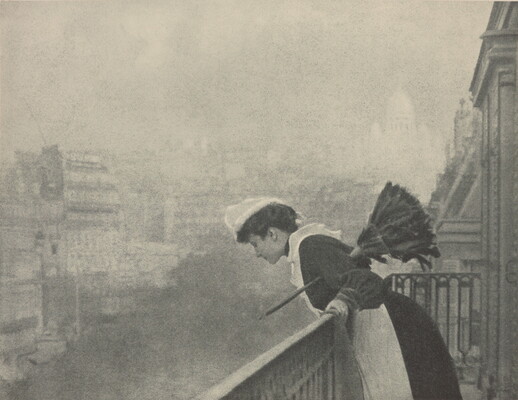 A black-and-white photograph of a White woman dressed as a maid in a white cap, black dress, and white pinafore holding a feather duster under her arm as she peers over a balcony at a smoggy cityscape.