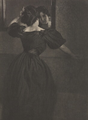 A black-and-white photograph of a young White woman, back to the camera, wearing a long, black dress with puffy sleeves fixing her hair in a mirror.