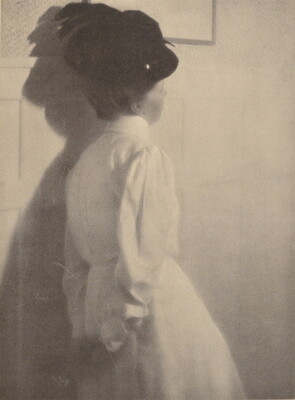 A black-and-white photograph of the back of a White woman wearing a black hat with a feather and a long-sleeved white dress, hands clasped behind her back.