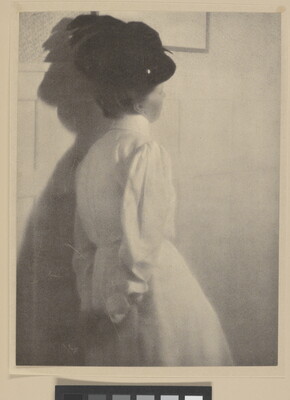 A black-and-white photograph of the back of a White woman wearing a black hat with a feather and a long-sleeved white dress, hands clasped behind her back.