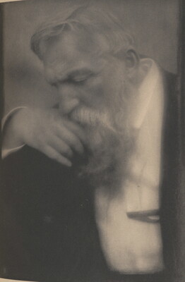 A black-and-white soft-focus photograph of an older White man with white hair and a long beard; he holds one hand up to his chin and looks down.