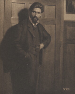 A sepia-toned portrait photograph of a White man with a beard in a three-piece suit, one hand in his pants pocket, leaning against a paneled wall.