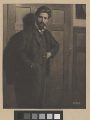 A sepia-toned portrait photograph of a White man with a beard in a three-piece suit, one hand in his pants pocket, leaning against a paneled wall.