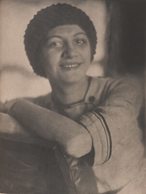 A black-and-white photograph of a smiling young White woman wearing a crocheted cap, one arm resting on what looks like the back of a chair.