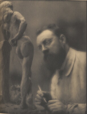 A sepia-toned photograph of a White man with glasses and a beard, holding a sculpting tool in his hand, looking a small sculpture of a nude woman.