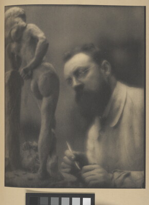 A sepia-toned photograph of a White man with glasses and a beard, holding a sculpting tool in his hand, looking a small sculpture of a nude woman.