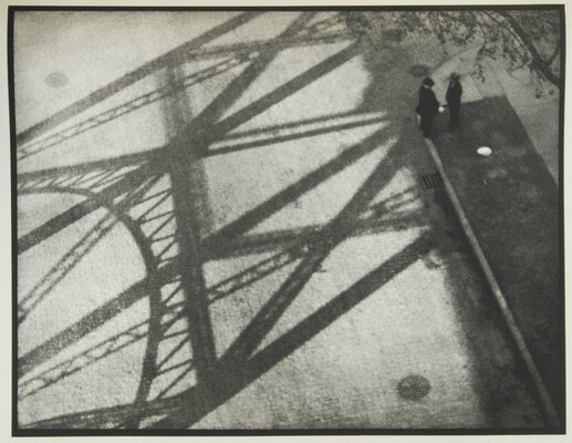 A black-and-white photograph taken from above of two people standing in the shadow of a bridge.