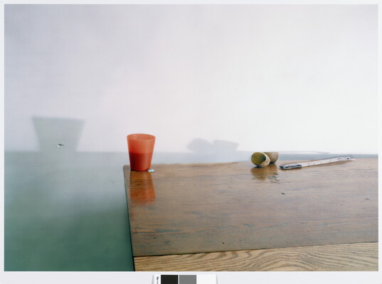 A color still life photograph of a plastic orange cup, a piece of fruit cut in half, and a flat piece of metal on a linoleum table casting shadows against a wall.