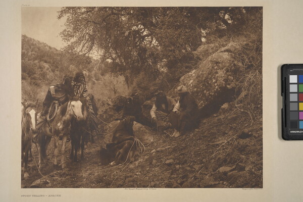 A sepia-toned photograph of a group of Indigenous people sitting on a grassy hill and horses.