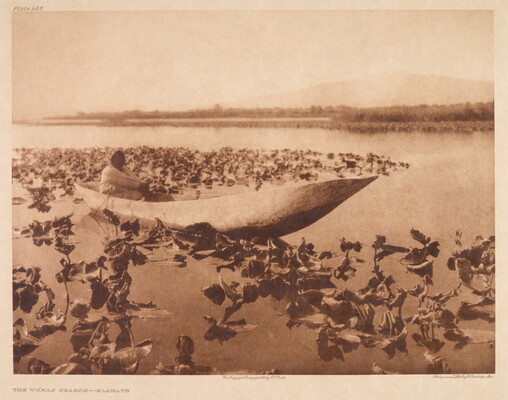A sepia-toned photograph of an Indigenous person sitting in a canoe on water surrounded by vegetation.