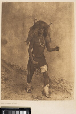 A sepia-toned photograph of an Indigenous man in cultural dress that includes a headpiece made of buffalo hide and horns.