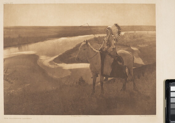 A sepia-toned photograph of a Native American man, in profile and wearing a full feathered headdress, on a horse looking off to a river in the background.